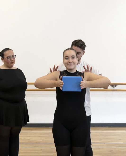 Dance instructor guiding a student holding a blue tablet, surrounded by fellow dance students watching attentively. Dance instructor guiding a student holding a blue tablet, surrounded by fellow dance students watching attentively.